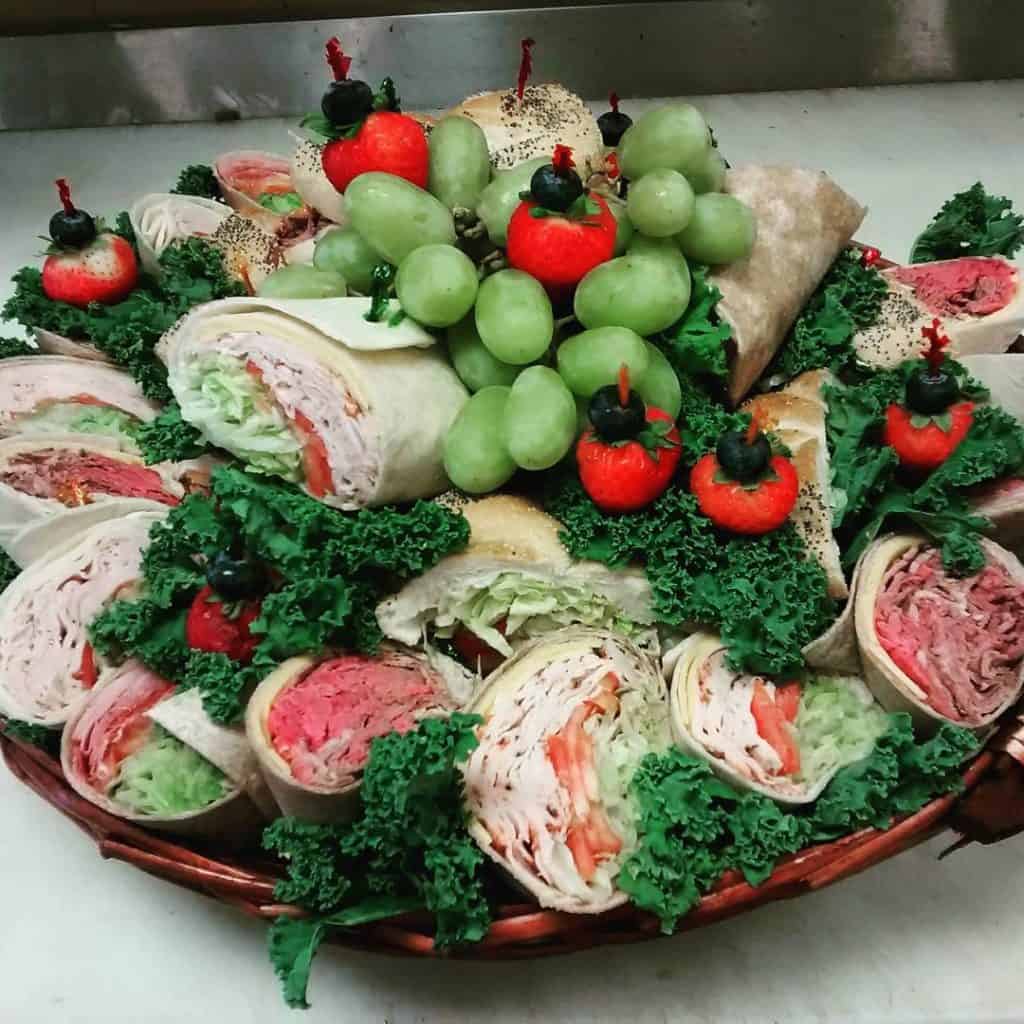 A platter filled with various wrap sandwiches containing lettuce, tomatoes, and meats. The wraps are garnished with leafy greens, clusters of green grapes, and decorative red vegetables resembling strawberries—a perfect offering from a catering company on Long Island, NY.