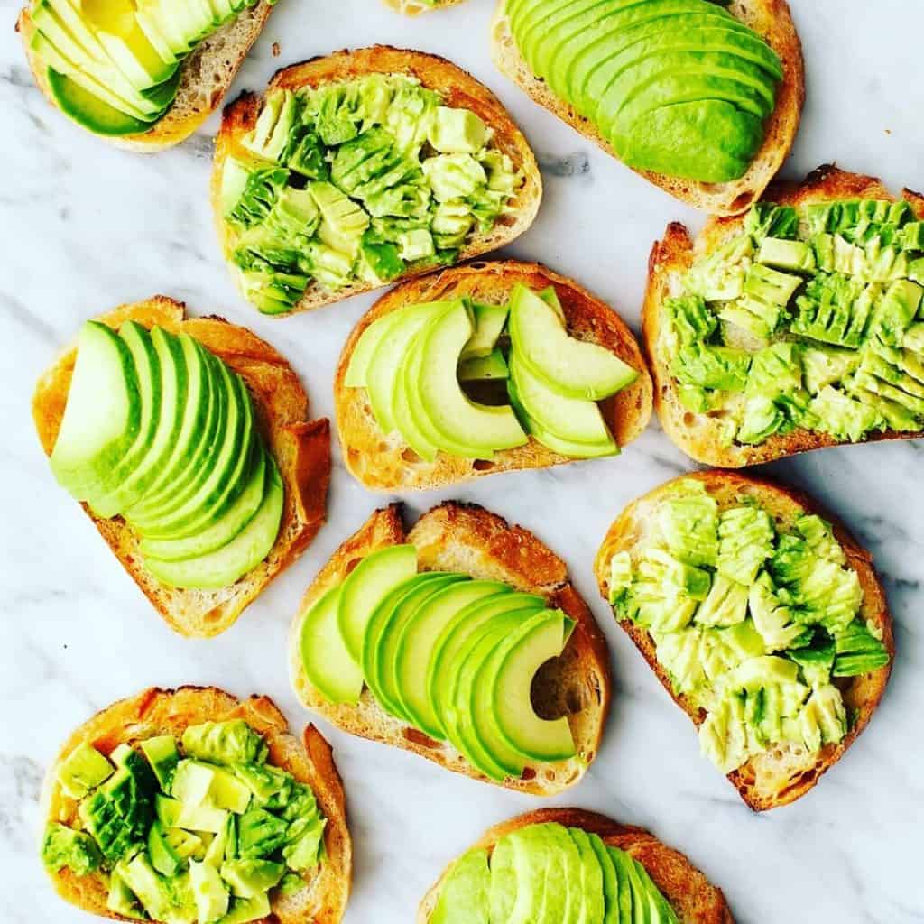 A variety of toast slices topped with sliced and mashed avocado are arranged on a marble surface. Perfect for events needing catering services Long Island NY, the avocado is spread in various styles, creating a visually appealing pattern of vibrant green hues.