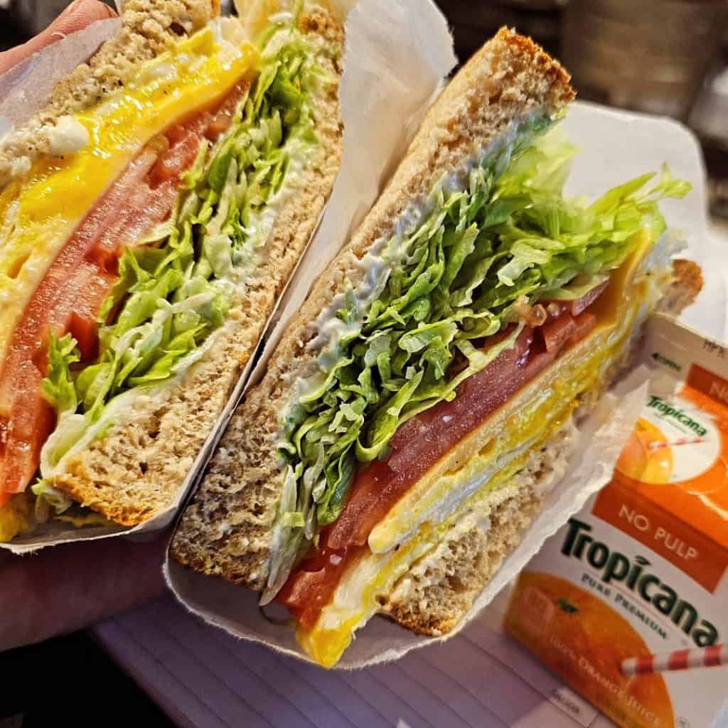 A close-up of a sandwich with lettuce, tomato, cheese, and mayonnaise on whole grain bread, cut in half. Next to it is a carton of Tropicana Pure Premium orange juice with a striped straw—perfectly presented by a premier catering company from Long Island NY.