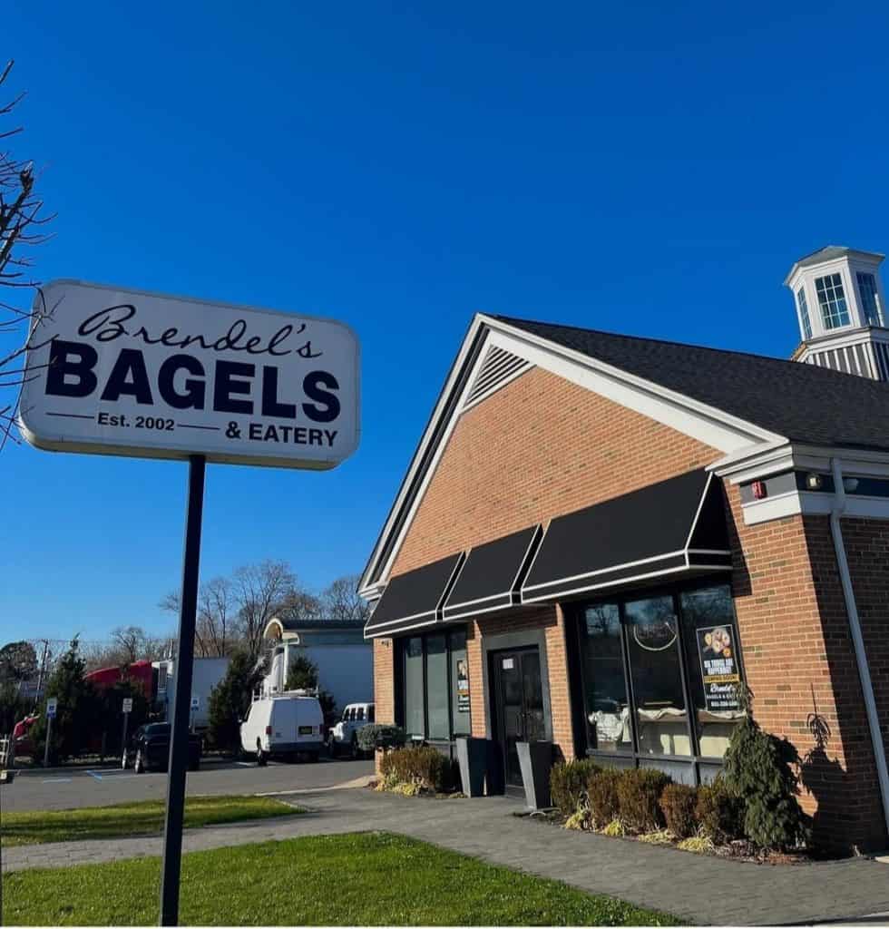 Exterior of Brendel's Bagels & Eatery with a brick facade and black awnings. The sign reads "Brendel's Bagels & Eatery Est. 2002." A white van, part of their catering company in Long Island, NY, is parked nearby under a clear blue sky.