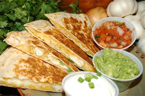 A plate of sliced quesadillas is arranged in a fan shape, with three small bowls containing sour cream, guacamole, and salsa. Fresh cilantro, onions, and garlic are visible in the background—a perfect presentation for catering services on Long Island NY.