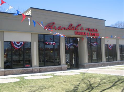 Storefront of Brendel's of New York Bagels & Eatery showcases large windows and a welcoming sign. Decorated with red, white, and blue bunting and flags, it proudly offers catering services in Long Island NY. The entrance is inviting with an "open" sign; grass and a sidewalk complete the scene.
