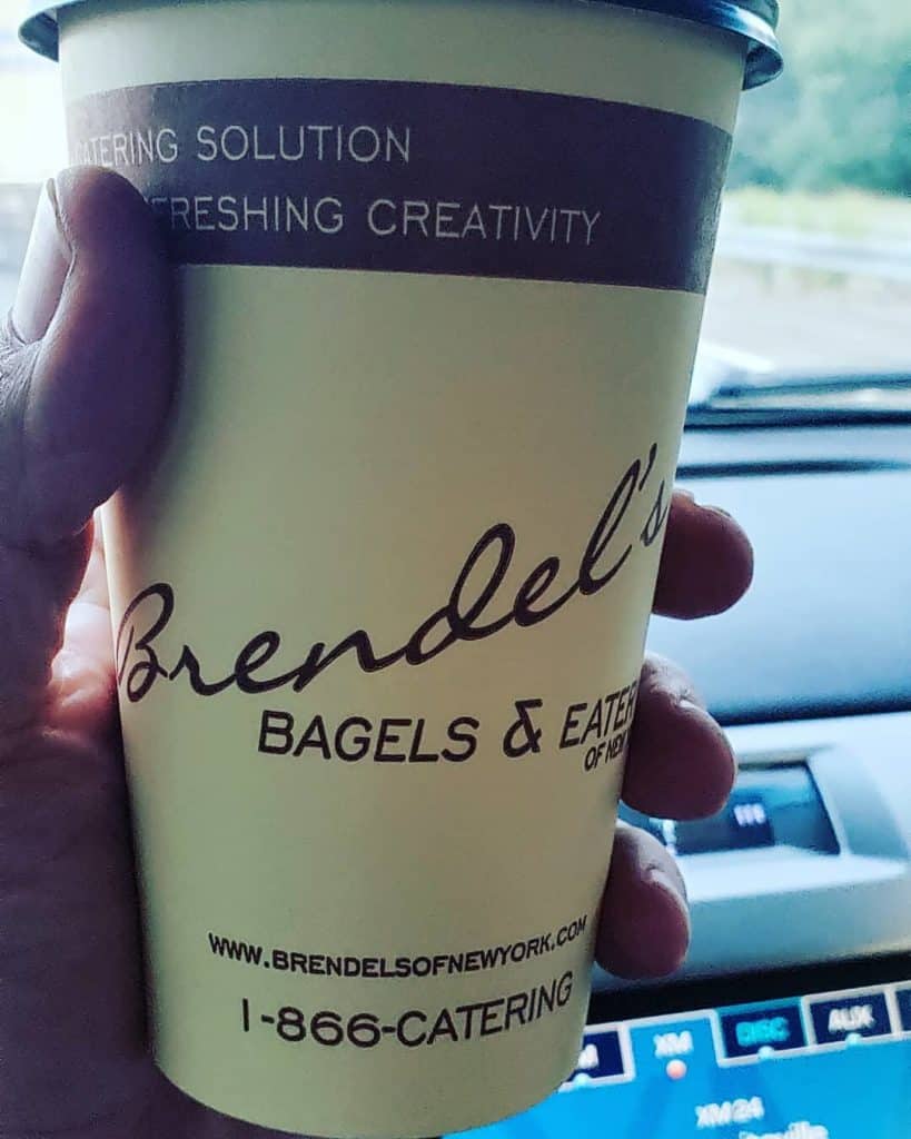 A hand holds a yellow coffee cup with a brown lid, emblazoned with "Brendel's Bagels & Eatery of New York" and contact information. As the car cruises through the roads, think about Brendel's trusted catering services Long Island NY for your next event.
