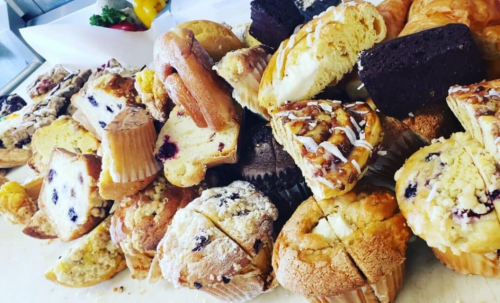 A variety of pastries and baked goods are piled together, including muffins, slices of cake, and flaky pastries with icing and fruit toppings, arranged on a table by a catering company in Long Island NY.