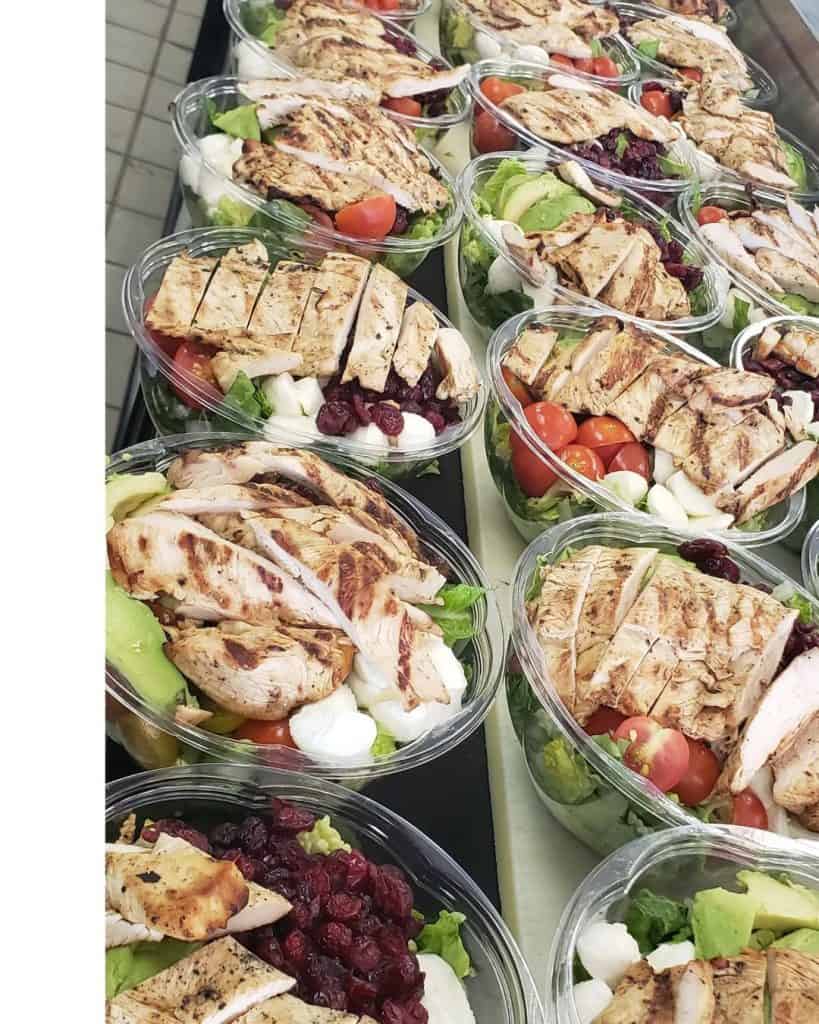 Multiple clear containers filled with salads, each topped with grilled chicken slices, are expertly prepared by our catering company in Long Island, NY. The salads include cherry tomatoes, mozzarella balls, leafy greens, avocado slices, and dried cranberries.