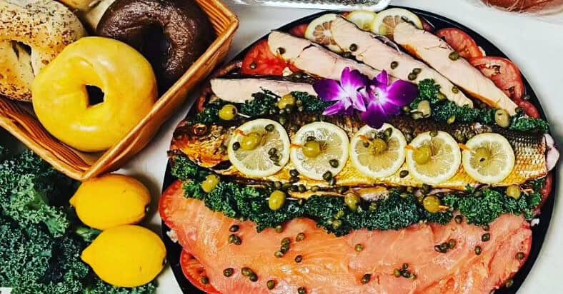 A platter featuring sliced smoked salmon, garnished with lemon slices, capers, and kale, with an ornamental purple flower on top. Nearby are assorted bagels and lemons in a basket—perfect for your next event with our catering services on Long Island NY.