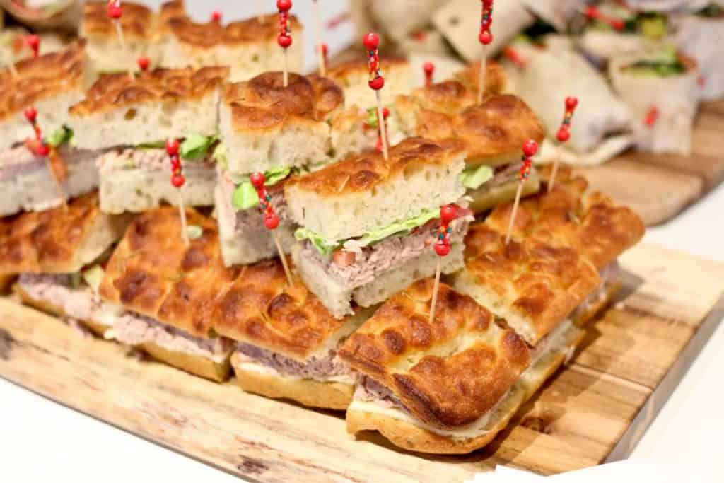A stack of focaccia sandwiches on a wooden board, each speared with red-topped toothpicks, showcases the delectable offerings from a top catering company in Long Island NY. Filled with crisp lettuce and savory slices of meat, these sandwiches are perfect for any event.