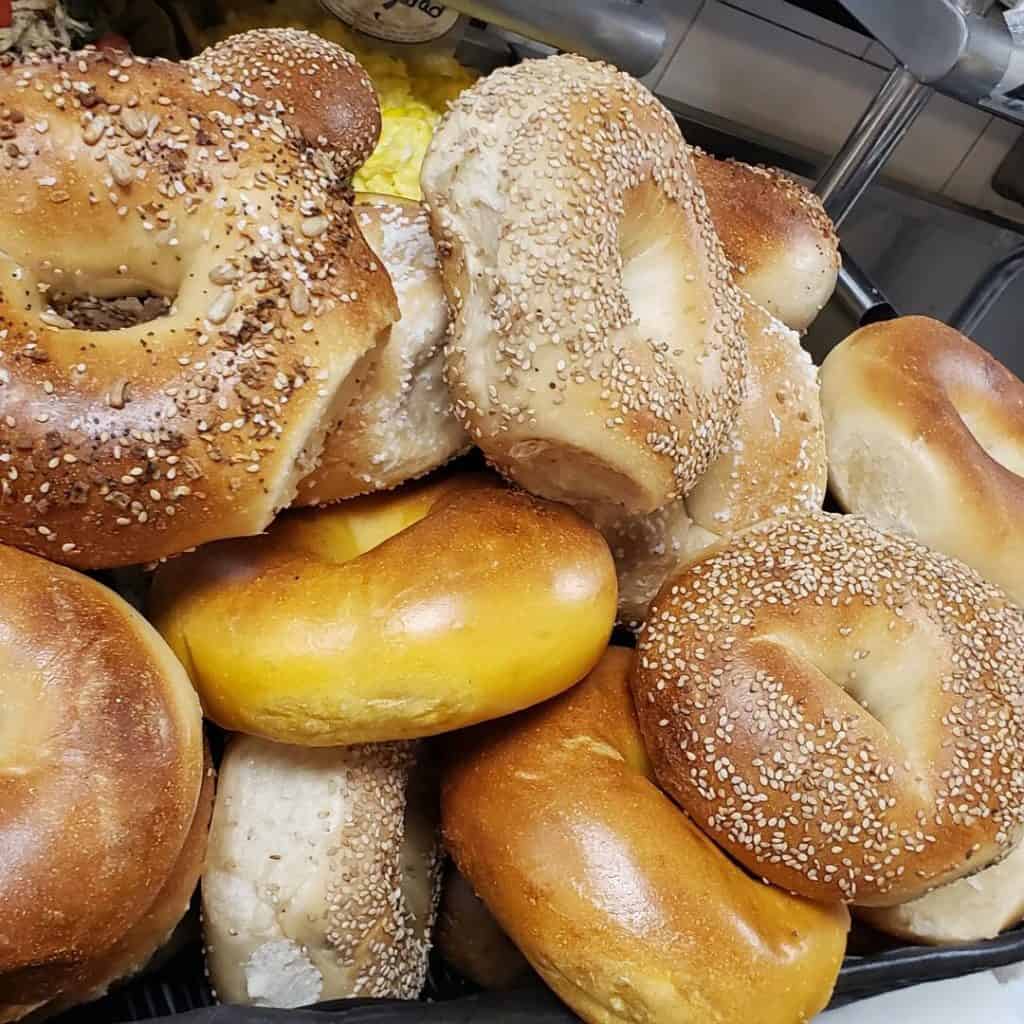 A pile of various bagels, including plain, sesame seed, and poppy seed, is artfully stacked by a premier catering company on Long Island, NY. Some bagels are golden brown while others are lighter in color, all displayed closely together to showcase their diverse textures and tempting toppings.