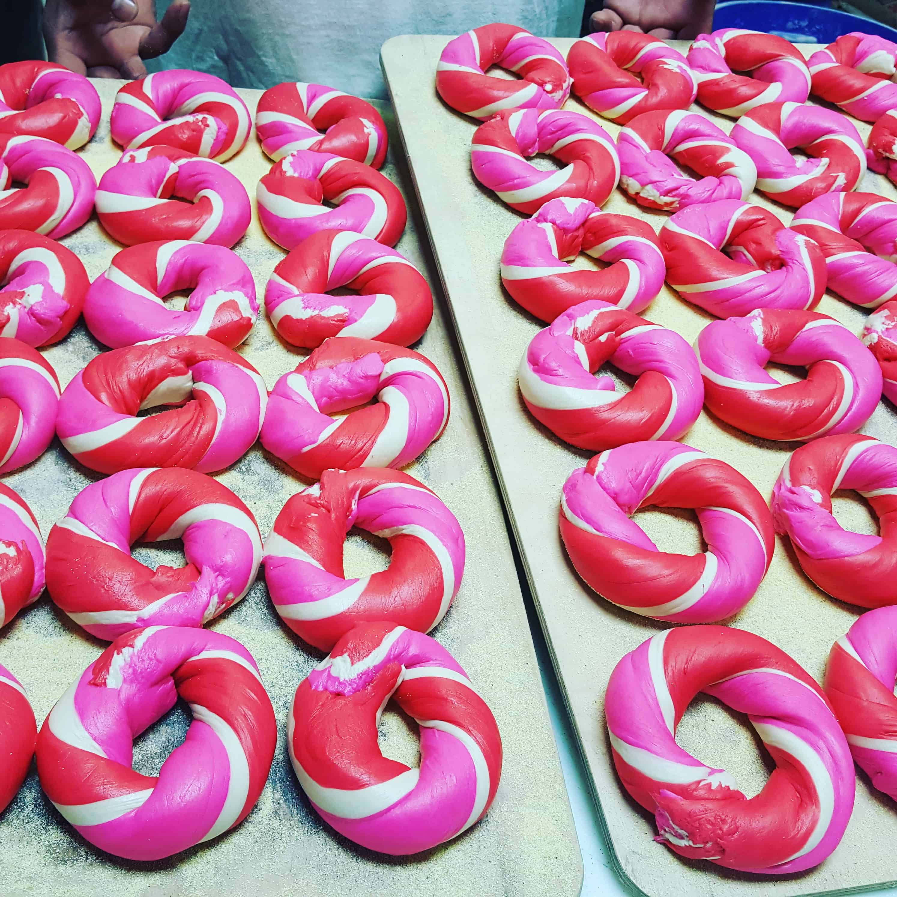 Several trays of red and pink swirled bagels are displayed, showcasing the unique touch of a catering company on Long Island, NY. Some bagels have noticeable gaps or imperfections, while a person's hands are visible at the top, suggesting involvement in their preparation.