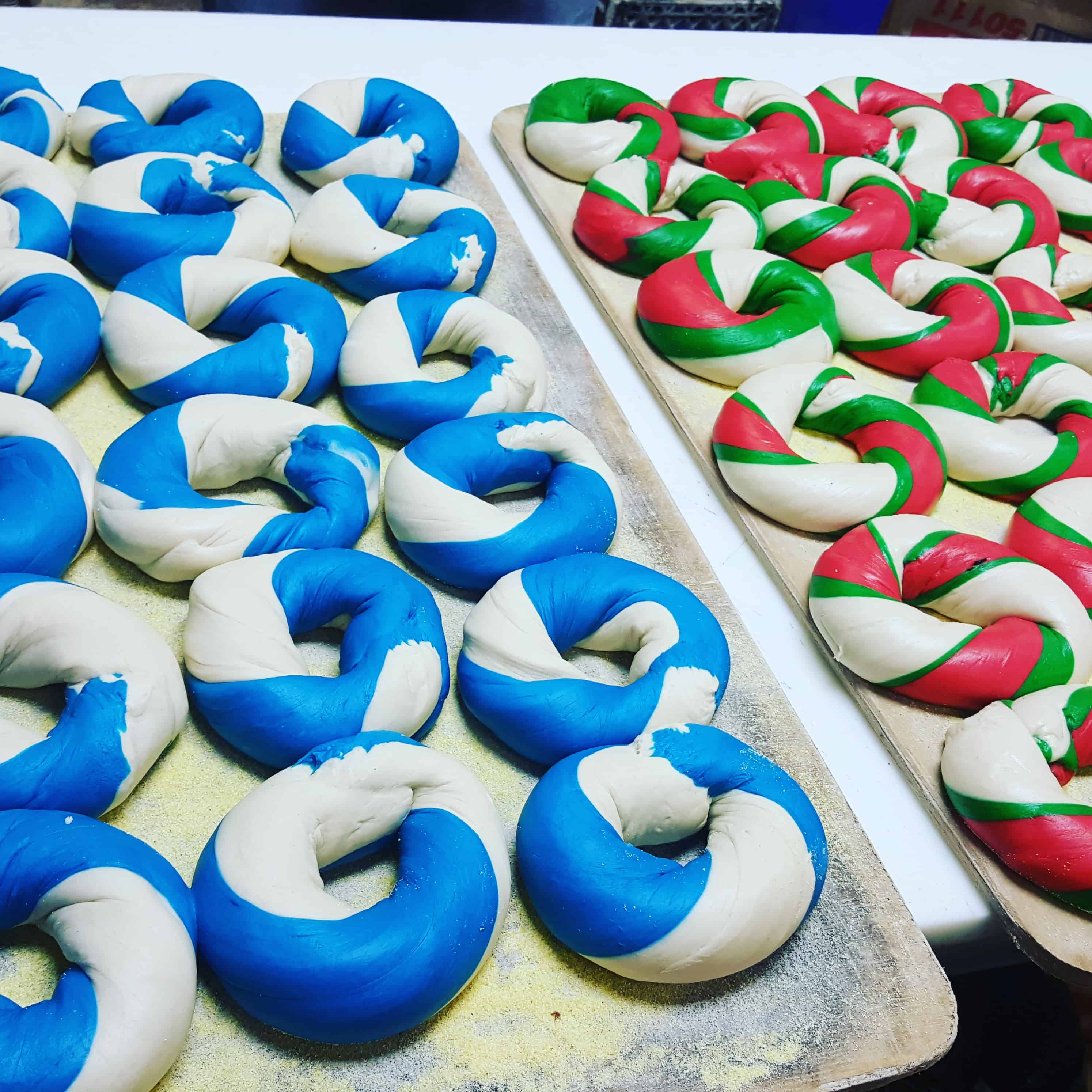 Two trays of unbaked bagels are shown, crafted by a top-notch catering company on Long Island NY. One tray boasts blue and white bagels, while the other displays red, green, and white. The colorful dough is twisted into a classic bagel shape, ready for baking perfection.