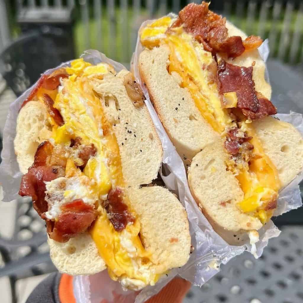 A hand holds two bagel halves filled with crispy bacon, melted cheese, and scrambled eggs. The bagels are sliced in half and wrapped in paper. Perfect for a brunch spread, these delights are ideal offerings from a catering company on Long Island, NY. A patio table is visible in the blurred background.