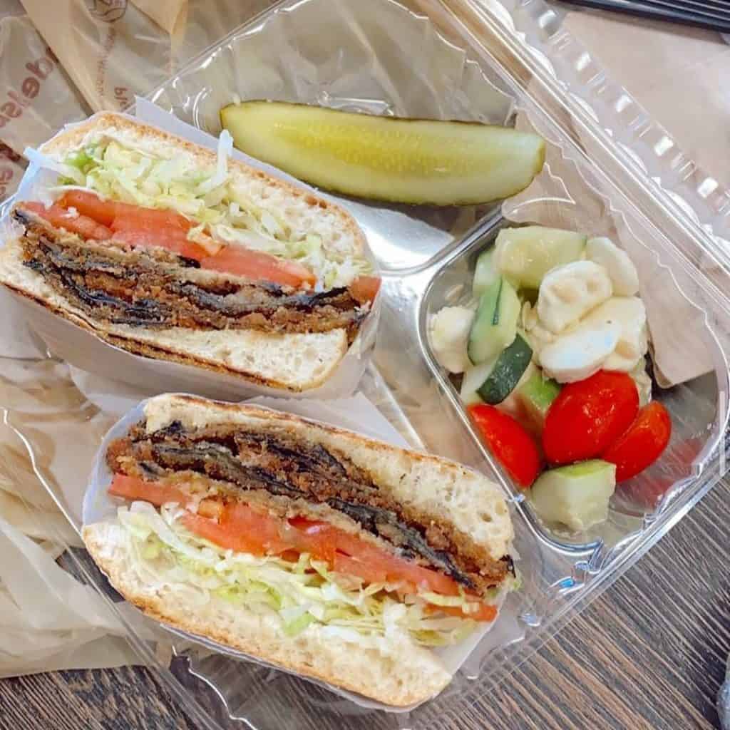 A clear plastic container, perfect for any catering services Long Island NY offers, holds a sandwich with lettuce, tomato, and a crispy patty—perhaps meat or veggie. It includes a dill pickle spear and a small salad with cucumber, cherry tomatoes, and mozzarella.