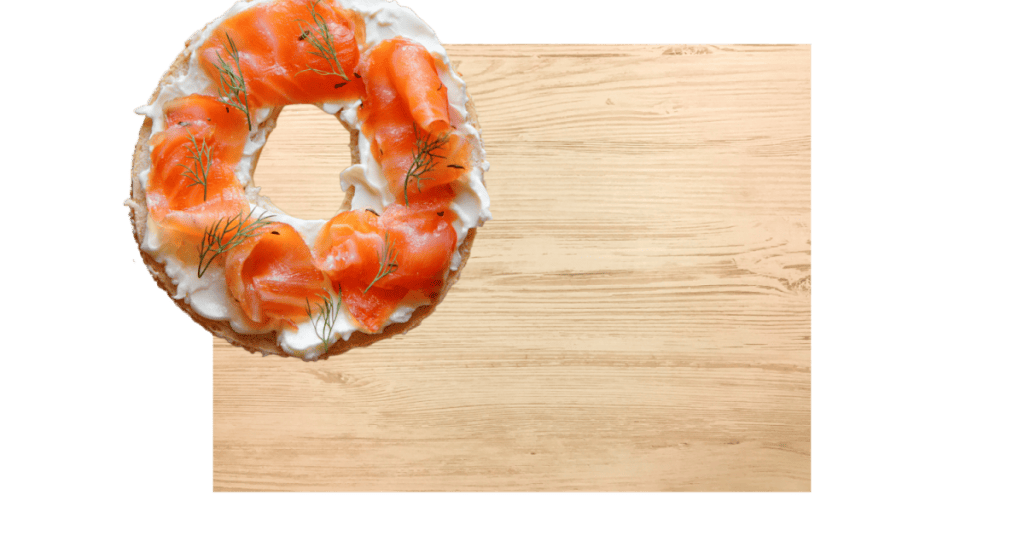 A bagel topped with cream cheese and slices of smoked salmon, garnished with dill, is artfully arranged on the top left corner of a wooden surface, showcasing the finesse of catering services Long Island NY.