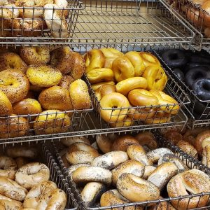 Wire baskets filled with various bagels, including poppy seed, plain, and charcoal varieties, are stacked on racks in a bakery display by a well-known catering company on Long Island, NY.