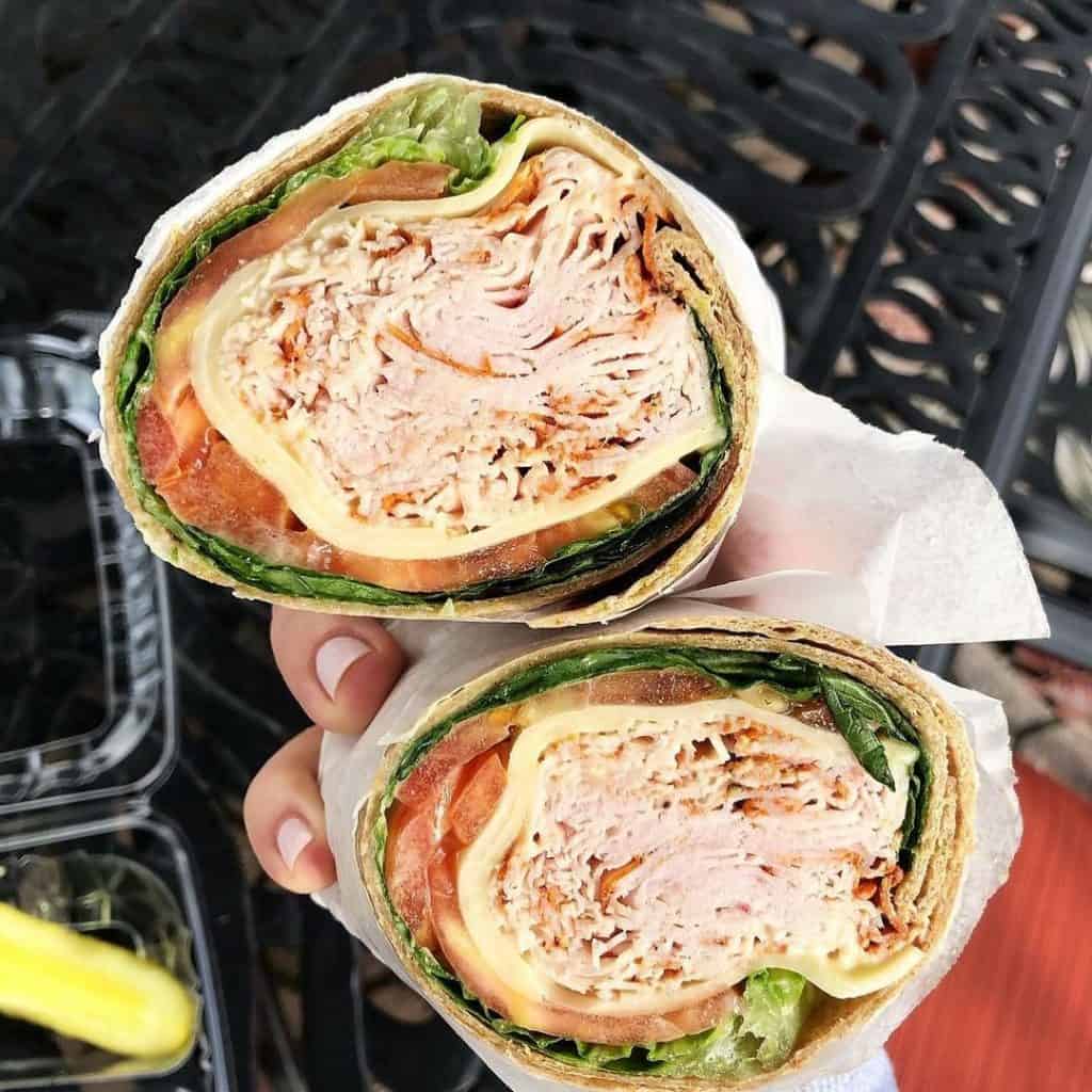 Two halves of a wrap sandwich filled with sliced turkey, cheese, lettuce, and tomato are wrapped in paper. The wrap is held by a hand with a pale pink manicure. A pickle sits in a plastic container on a black metal table—a delightful option from catering services Long Island NY.