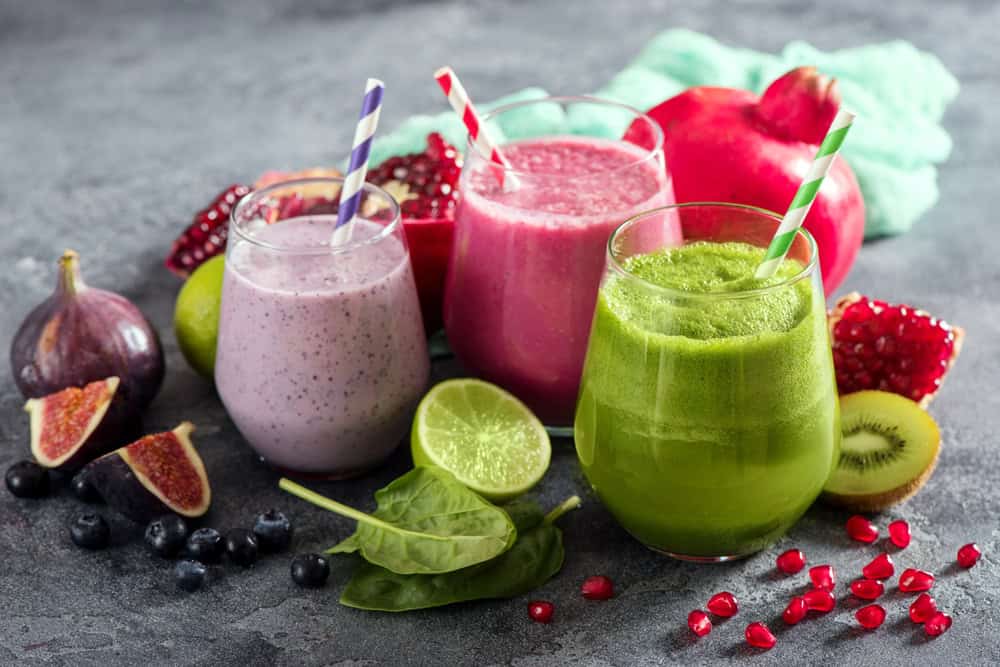 Three smoothies in glasses with straws: a vibrant purple with blueberries and figs, a lively pink with pomegranate and lime, and a refreshing green with kiwi and spinach. Surrounded by fresh fruits on a gray surface, perfect for any event organized by catering services on Long Island, NY.