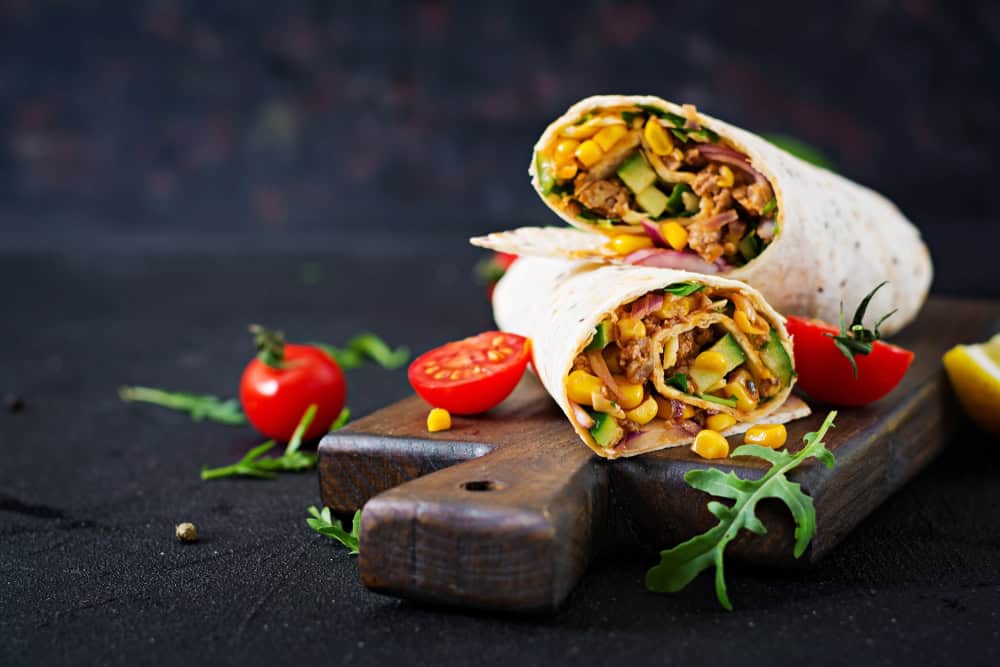 A wooden board presents two halves of a wrap bursting with colorful ingredients like corn, lettuce, and sliced meat. Fresh cherry tomatoes, arugula leaves, and a lemon wedge surround the board—a perfect showcase of the exquisite offerings from a catering company in Long Island NY.