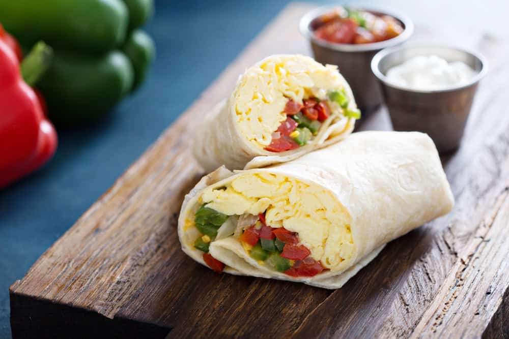 A breakfast burrito cut in half, filled with scrambled eggs, green peppers, and tomatoes, is placed on a wooden board. Two small metal cups containing salsa and sour cream are beside it. In the background, vibrant bell peppers add color—just a taste of what our catering services Long Island NY can offer.