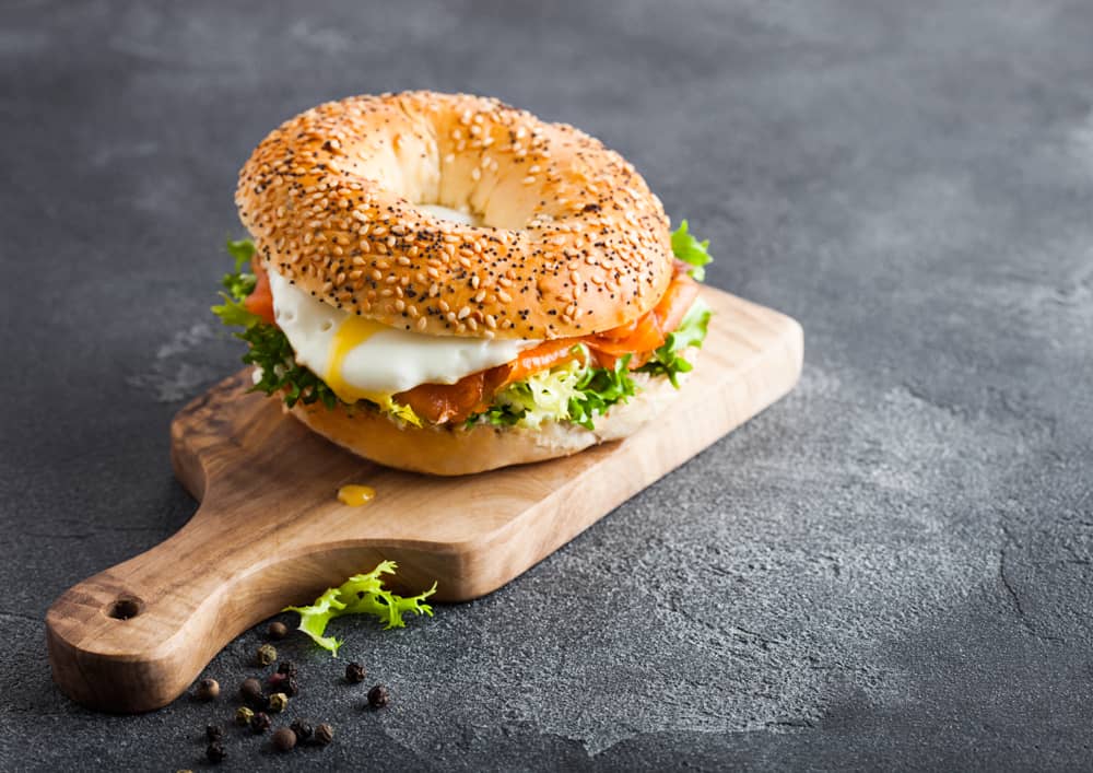 A sesame seed bagel sandwich with smoked salmon, cream cheese, and lettuce rests on a wooden board against a dark textured background sprinkled with peppercorns, perfect for showcasing the artisanal touch of a catering company Long Island NY.
