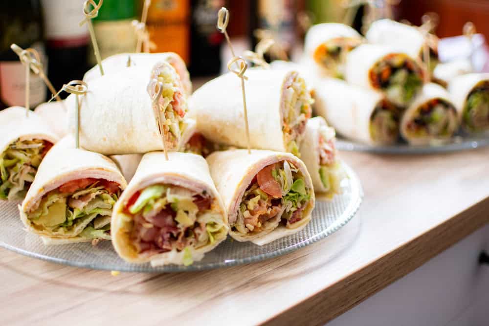 Close-up of assorted wraps on a plate, perfect for catering services in Long Island NY. The wraps are sliced and filled with lettuce, tomatoes, and various meats, secured with toothpicks. Another plate of wraps is visible in the background on a wooden surface.