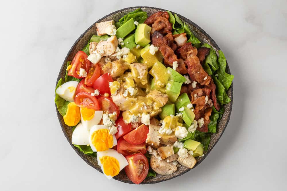 A Cobb salad on a plate with chopped lettuce, grilled chicken, avocado, bacon, hard-boiled eggs, cherry tomatoes, and blue cheese crumbles is drizzled with dressing—perfect for any catering event by the top catering company on Long Island NY.