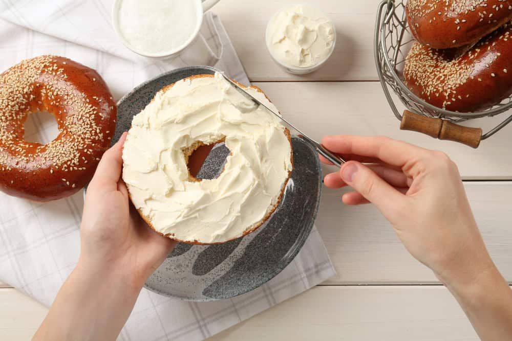 A hand spreads cream cheese on a sesame bagel over a dark plate. Nearby, more bagels await in a wire basket beside a bowl of cream cheese on a checkered cloth. This enticing scene is part of the delicious offerings from a premier catering company in Long Island, NY.
