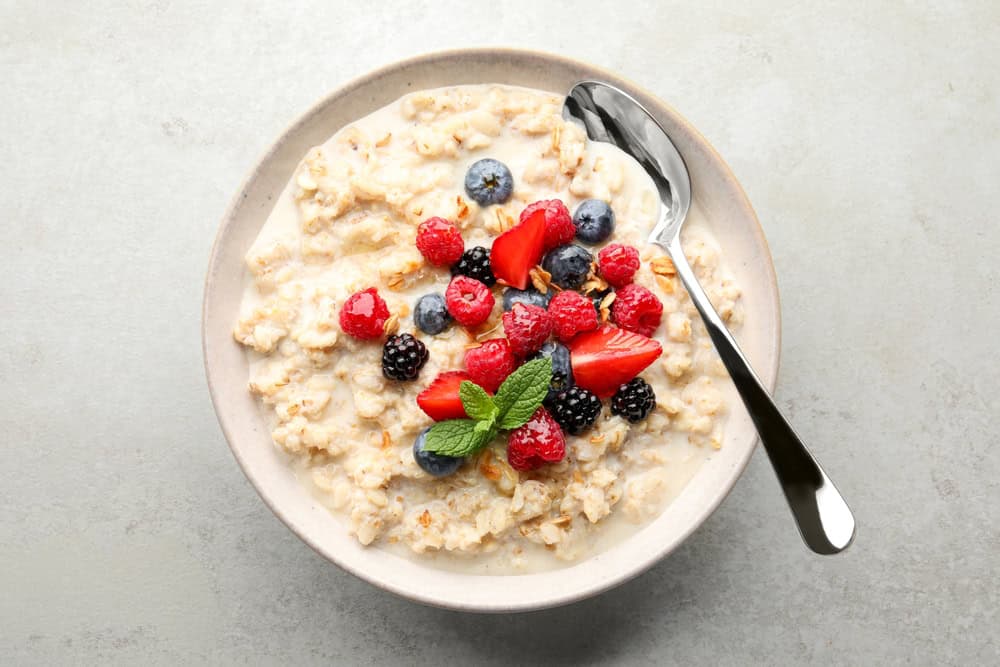 A bowl of oatmeal topped with fresh strawberries, raspberries, blueberries, and blackberries is artfully presented by a renowned catering company Long Island NY. A sprig of mint garnishes the bowl, while a spoon rests inside on a light gray surface.