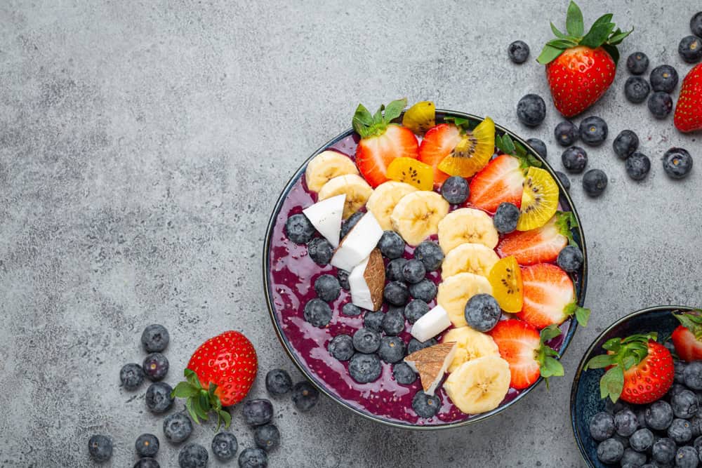 A vibrant smoothie bowl topped with sliced bananas, strawberries, kiwi, blueberries, and coconut chunks graces the gray textured background. Scattered berries and a peek of another bowl hint at the exquisite offerings available from a top catering company on Long Island, NY.