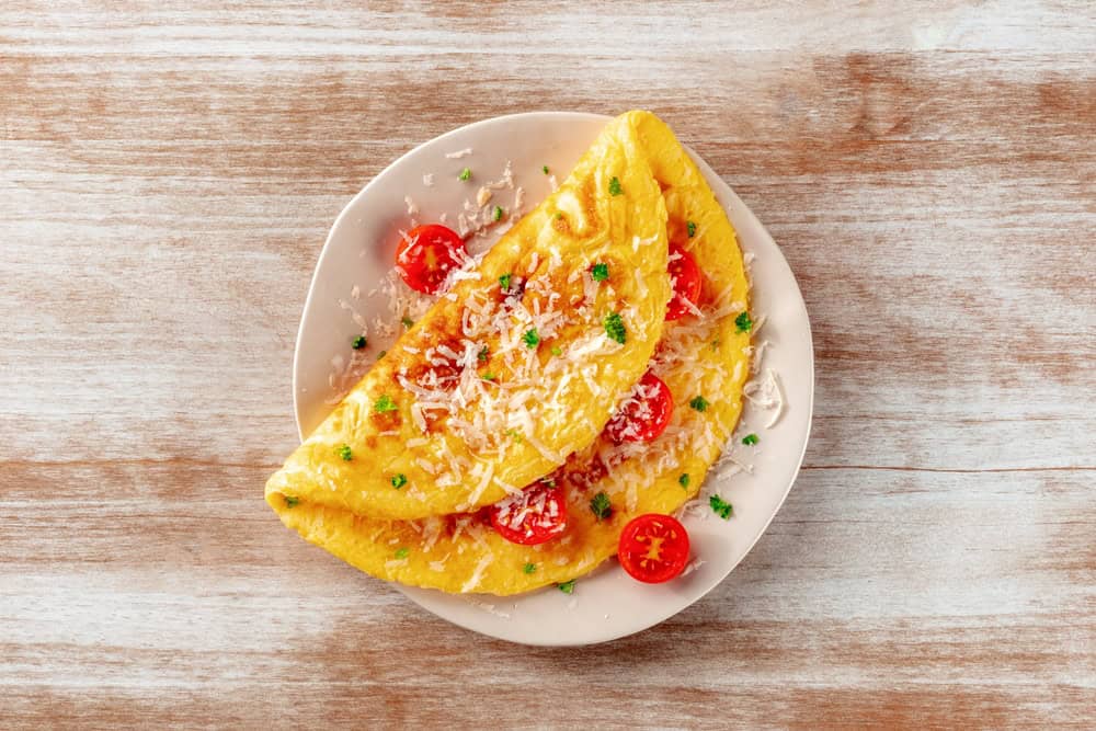 A fluffy omelette filled with cherry tomatoes and garnished with grated cheese and herbs, served on a white plate. This delightful dish, crafted by a catering company on Long Island, NY, rests invitingly on a rustic wooden table.