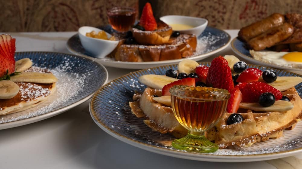 On a table, delightful breakfast plates await: waffles crowned with strawberries, blueberries, and bananas; pancakes graced with bananas and strawberries; a glass of syrup nearby; eggs with sausages tempting in the background. Perfectly crafted by a top catering company on Long Island, NY.