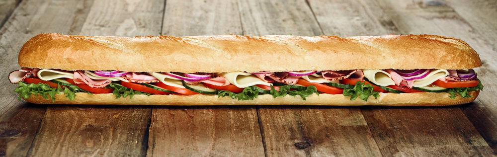 A long submarine sandwich rests on a wooden surface, filled with layers of lettuce, sliced tomatoes, cucumbers, cheese, deli meats, and red onion slices—a perfect offering from top catering services in Long Island NY.