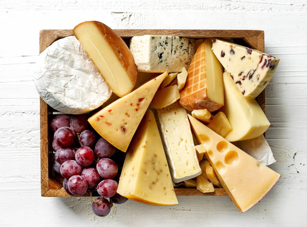 A wooden tray from a catering company in Long Island, NY, offers an assortment of cheeses in various textures and colors, accented by red grapes on the left. The scene is set against a white wooden surface, perfect for elevating any occasion.