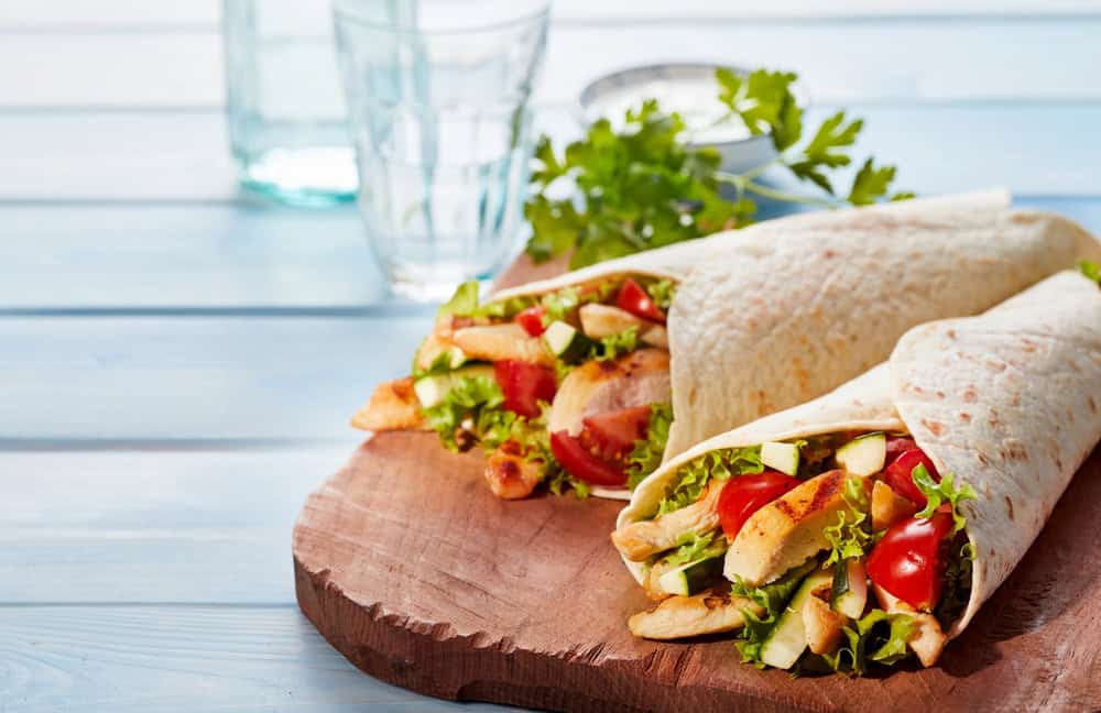 Two chicken wraps with lettuce, tomatoes, and other fresh vegetables are arranged on a wooden board. A sprig of parsley and a small bowl of sauce linger in the background, alongside two empty glasses on a blue wooden surface—perfect for showcasing at any catering event in Long Island NY.