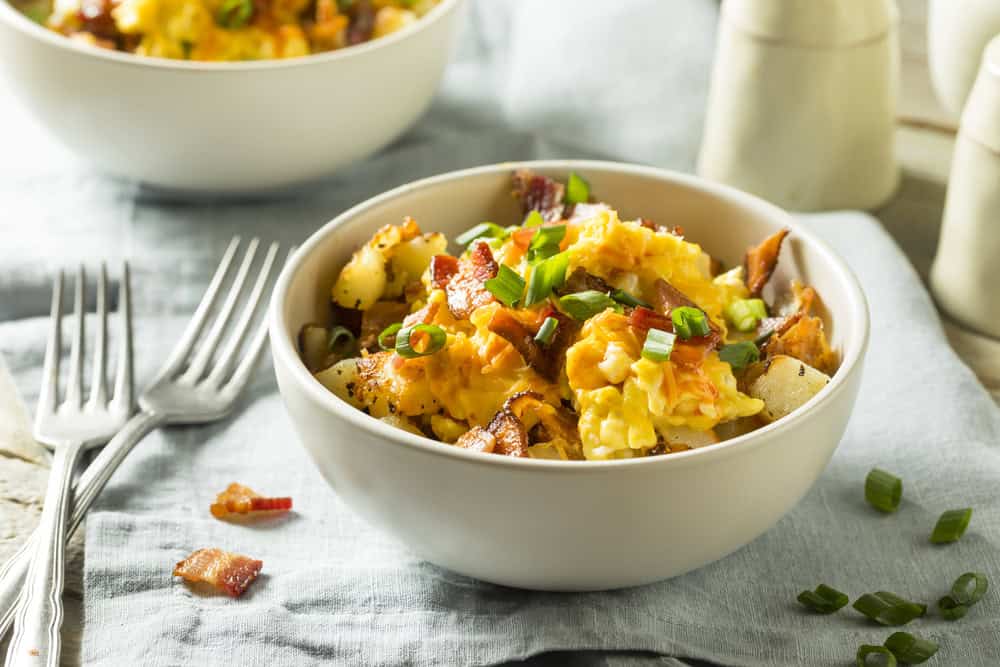 A bowl filled with scrambled eggs, crispy bacon, diced potatoes, and chopped green onions beckons breakfast lovers. Perfectly poised for your mornings or events, this dish showcases the excellence of catering services Long Island NY has to offer, with two forks resting on a light blue cloth nearby.